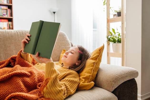 Smiling girl reading book and lying with knitted blanket on sofa at home model released, Symbolfoto property released, ELMF01079