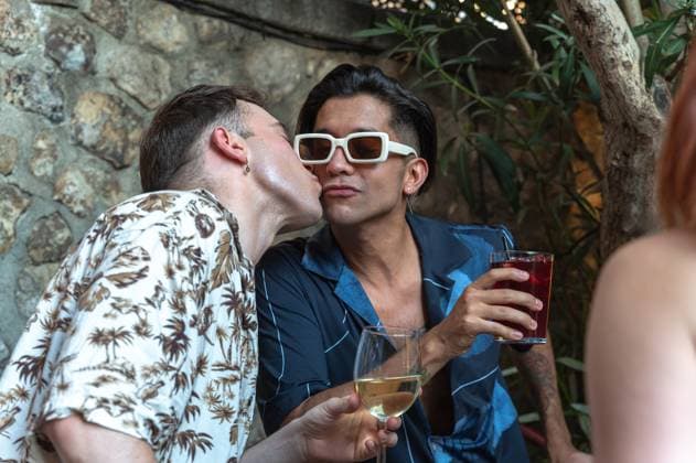 IMAGO Creative, Lifestyle, Feste im Garten #2 A tender scene captures one man kissing another on the cheek while holding glasses of wine at a lively garden party, reflecting a moment of love and celebration. Model Released DavidPrado_ID25521_737856_147 Copyright: xDavidxPradox
