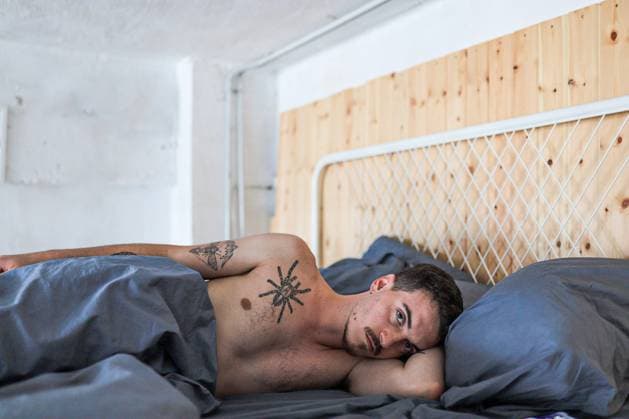 Creative Highlights Symbolbilder 783449156 Young man with tattoos lying on his side in bed with a thoughtful expression. The room has wooden elements and gray bedding, creating a cozy, relaxed atmosphere. Model Released Gaorstudio_ID23045_670157_003 Copyright: xGaorstudiox RECORD DATE NOT STATED