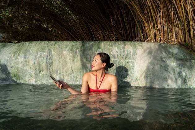 An asian woman in a red swimsuit enjoys a peaceful soak at Terme di Saturnia, Tuscany, Italy She holds a phone, surrounded by natural beauty and calming waters Model Released JuanAlbertoRuiz_ID25009_724433_003 Copyright: xJuanxAlbertoxRuizx RECORD DATE NOT STATED