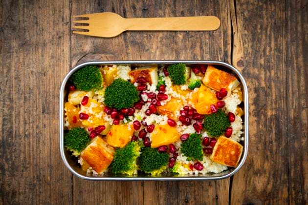 Stainless steel lunch box with couscous, broccoli, halloumi, oranges, chili dressing, and pomegranate seeds, on a wooden table with a wooden fork., LVF09377