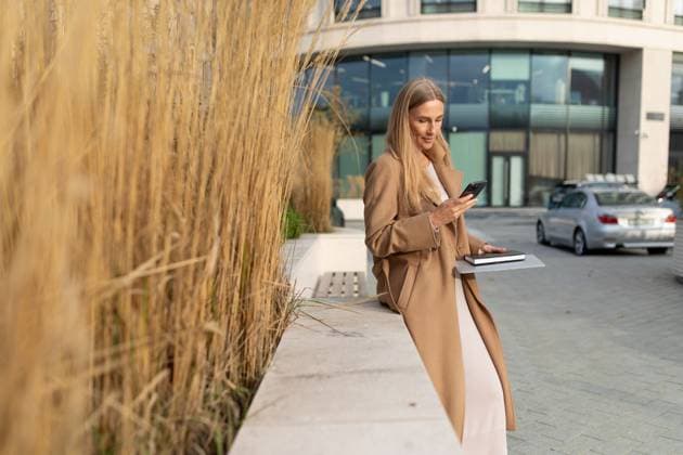 Creative Highlights Symbolfotos Eine Geschäftsfrau benutzt ihr Telefon, während sie sich vor einem Geschäftszentrum an eine Wand lehnt A businesswoman with long blonde hair is wearing a beige coat. She is standing outside a business center and using her phone. She has a notebook in her other hand. ,model released, Symbolfoto Copyright: xZoonar.com IvanxTraimakx 22428399 ,model released, Symbolfoto ,property released