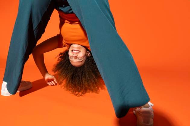 Creative Highlights Symbolbilder Young woman with eyes closed looking through legs in front of orange background model released, Symbolfoto, MIKF00534
