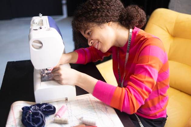 Junge afrikanische Frau an der Nähmaschine. Charmantes schwarzes Mädchen in rotem Pullover Young African woman working at sewing machine. Charming black girl in red sweater with measuring tape. Materials for sewing and fabric patterns in workplace. Seamstress is engaged in needlework ,model released, Symbolfoto Copyright: xZoonar.com DashaxPetrenkox 22600950 ,model released, Symbolfoto ,property released