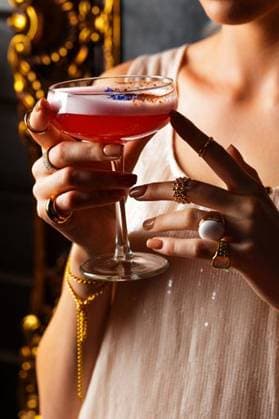 Creative Highlights Symbolbilder Elegant woman holding a cocktail in a coupe glass, adorned with gold jewelry, in a dimly lit setting. 28318433 ,model released, Symbolfoto RECORD DATE NOT STATED