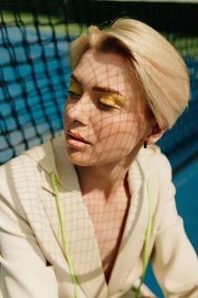 Creative Highlights Symbolbilder A stylish young adult with blonde hair and vibrant yellow eyeshadow posing by a tennis court net, casting shadows on their face. 28318438 ,model released, Symbolfoto RECORD DATE NOT STATED