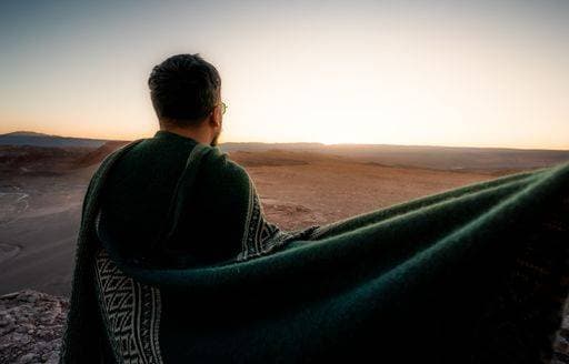Creative Highlights Symbolbilder A man wrapped in a green blanket gazes at a serene desert landscape during sunset, capturing a moment of solitude and reflection. 26748219 ,model released, Symbolfoto RECORD DATE NOT STATED
