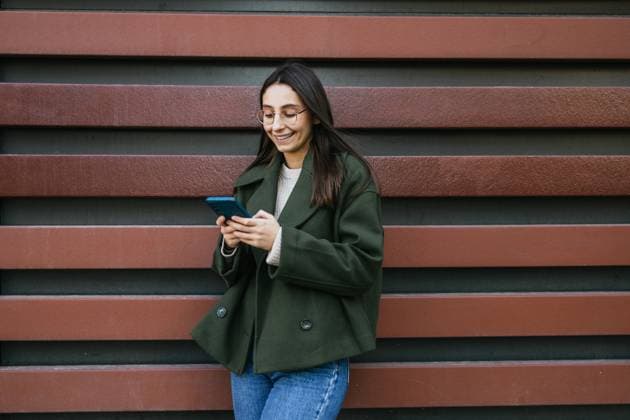 Creative Highlights Symbolbilder A woman in a green coat is smiling while using her smartphone She stands against a decorative striped wall, creating a casual yet stylish urban scene Model Released DavidMunoz_ID25897_748130_023 Copyright: xDavidxMunozx