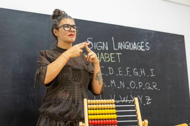 An instructor teaching sign language in a classroom, standing in front of a chalkboard with the alphabet written on it An abacus is visible in the foreground Model Released JuanAizpuru_ID25565_738874_008 Copyright: xJuanxAizpurux