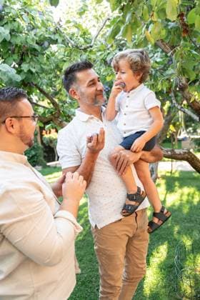 IMAGO Creative, Jahreszeit Sommer, Food, Pflaume A joyful gay couple shares a sweet moment with their young son in a lush garden. The child is smiling and eating plums, surrounded by greenery and natural light Model Released ManuelRuiz_ID25867_747165_012 Copyright: xManuelxRuizx RECORD DATE NOT STATED