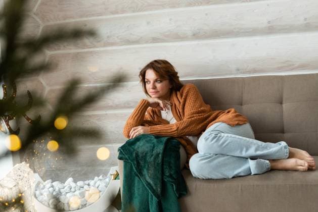 A woman sits cozily on a couch, adorned in a warm orange sweater and light blue jeans, gently looking away while surrounded by Christmas decorations and soft, festive lights. Model Released Shapovalphoto_ID25696_741677_014 Copyright: xShapovalphotox RECORD DATE NOT STATED