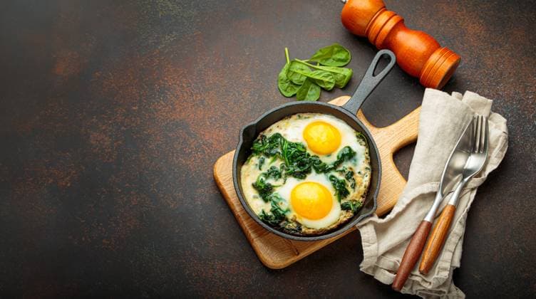 Gesundes Spinat-Omelett in gusseiserner Pfanne zum Frühstück Ansicht von oben Two fried eggs with spinach in a cast iron pan on a wooden board with fresh ingredients, healthy breakfast Copyright: xZoonar.com OlenaxYeromenkox 22625955