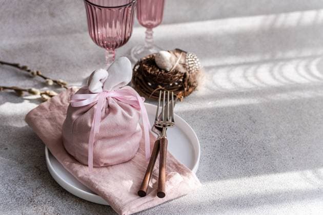 An artistic Easter table arrangement featuring a bunny-themed pink cloth wrap, elegantly tied, accompanied by unique pink glasses and a delicate willow nest containing speckled eggs, all set against a backdrop of soft light and shadows. Model Released AnnaBogush_ID26004_750158_003 Copyright: xAnnaxBogushx RECORD DATE NOT STATED
