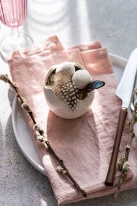 From above, an artistic Easter table arrangement features a porcelain bowl with speckled eggs, adorned with delicate feathers, beside a pastel pink napkin and elegant silverware under soft daylight. Model Released AnnaBogush_ID26004_750160_005 Copyright: xAnnaxBogushx RECORD DATE NOT STATED