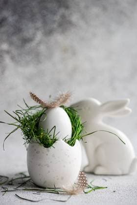 A meticulously crafted Easter setting featuring a white speckled egg cradling a nest of fresh green grass, topped with a delicate feather, with a sleek white ceramic bunny nearby, set against a soft, textured gray background. Model Released AnnaBogush_ID26004_750188_017 Copyright: xAnnaxBogushx RECORD DATE NOT STATED