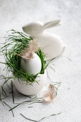 A meticulously styled Easter setting featuring a white egg cradled in vibrant green grass, delicately accented with soft feathers, beside a sleek ceramic bunny on a textured surface. Model Released AnnaBogush_ID26004_750189_018 Copyright: xAnnaxBogushx RECORD DATE NOT STATED