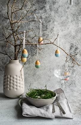 An Easter table setting featuring a ceramic vase with suspended painted eggs from bare tree branches and a bowl of grass accented by subtle textures of a tablecloth Model Released AnnaBogush_ID26004_750202_022 Copyright: xAnnaxBogushx RECORD DATE NOT STATED