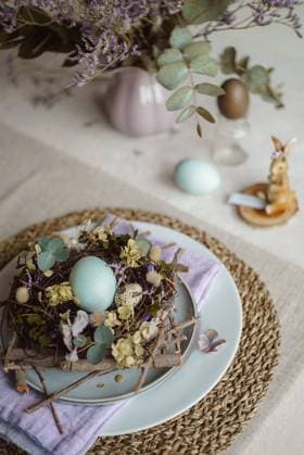 Top view of an Easter table setting featuring a plate with a rustic nest of twigs and naturally dyed eggs, surrounded by floral decorations and subtle tableware accents under soft, natural lighting. Model Released CamilaMacedo_ID26016_750688_002 Copyright: xCamilaxMacedox RECORD DATE NOT STATED