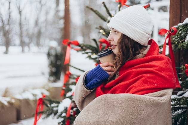 Young woman wrapped in a red scarf, holding a coffee cup, and smiling, seated near a festive Christmas tree on a snowy day. Model Released HannaYakimenko_ID26106_753105_020 Copyright: xHannaxYakimenkox