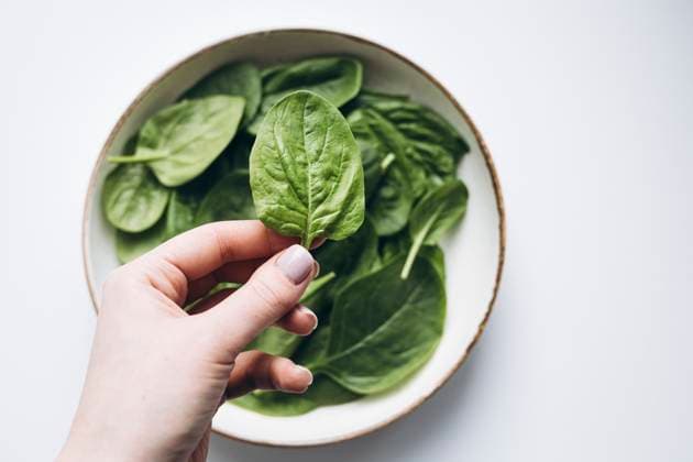 A hand holding a fresh green spinach leaf over a bowl filled with more spinach leaves. The vibrant green color emphasizes freshness and health, ideal for culinary themes. Model Released HannaYakimenko_ID26198_755897_005 Copyright: xHannaxYakimenkox