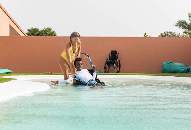 A woman assists a black boy in a wheelchair with cerebral palsy to enjoy the pool, promoting inclusivity and accessibility. The image highlights joy and companionship in an outdoor setting. Model Released JuanAizpuru_ID26084_752375_018 Copyright: xJuanxAizpurux