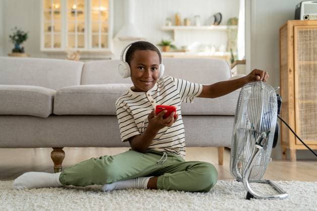IMAGO Creative, Jahreszeit Sommer, Hitze Lächelnder, glücklicher afroamerikanischer Junge, der sich zu Hause vor einem elektrischen Ventilator entspannt Smiling happy African American boy child wearing headphones listening music or watching internet video while relaxing at home in front of electric fan. Children and summer heat ,model released, Symbolfoto Copyright: xZoonar.com DmitriixMarchenkox 22690747 ,model released, Symbolfoto ,property released