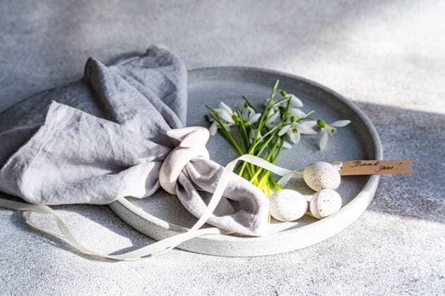 A plate elegantly displays spring snowdrops alongside speckled Easter eggs, a grey linen cloth, and a Thank You tag, all set on a textured surface. Model Released AnnaBogush_ID26489_764292_003 Copyright: xAnnaxBogushx