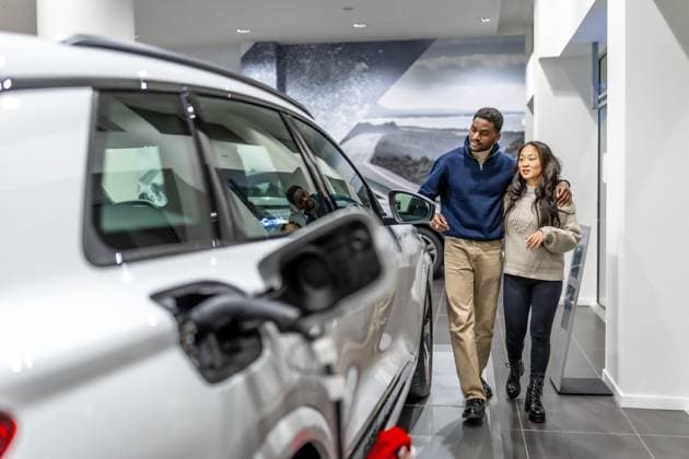Ein multiethnisches Paar geht durch ein Autohaus und wählt fröhlich sein neues Elektrofahrzeug aus, während es den modernen Ausstellungsraum genießt Multiethnic couple walking through a car dealership, happily selecting their new electric vehicle while enjoying the modern showroom experience Copyright: imageBROKER UnaixHuizi ibxuhu12460353.jpg ,model released, Symbolfoto ,property released
