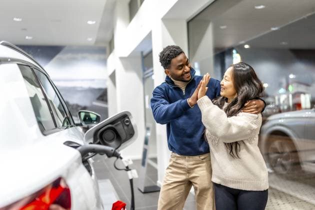 Ein glückliches multiethnisches Paar feiert den Kauf seines neuen Elektroautos mit einem High Five im Autohaus und strahlt vor Freude und Begeisterung Happy multiethnic couple celebrating their new electric car purchase with a high five at the dealership, radiating joy and excitement Copyright: imageBROKER UnaixHuizi ibxuhu12460363.jpg ,model released, Symbolfoto ,property released