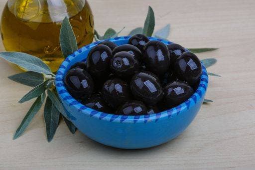 A blue bowl filled with black olives next to a bottle of olive oil and olive leaves on a wooden table. 25070648 RECORD DATE NOT STATED