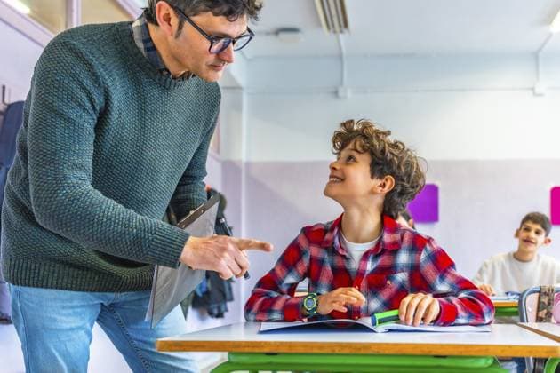 Grundschullehrer, der sich mit einem lächelnden Schüler unterhält, während er an seinem Schreibtisch in einem fröhlichen Klassenzimmer eine Lektion erklärt Elementary school teacher engaging with a smiling student while explaining a lesson at their desk in a cheerful classroom setting Copyright: imageBROKER UnaixHuizi ibxuhu12678454.jpg ,model released, Symbolfoto ,property released