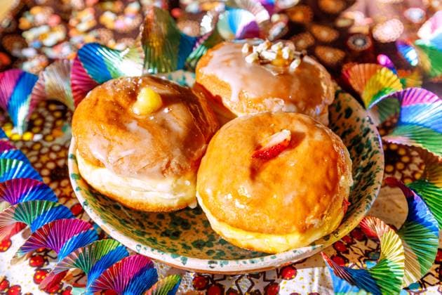 bunte gefüllte Faschingskrapfen mit Zuckerguss (Erdbeer-Limes, Eierlikör, weiße Schokoladencreme), Keramikteller weiß-grün melliert, gemusterte Tischdecke, bunte Dekospiralen *** colorful filled carnival doughnuts with icing strawberry lime, egg liqueur, white chocolate cream, ceramic plate white green mottled, patterned tablecloth, colorful decorative spirals