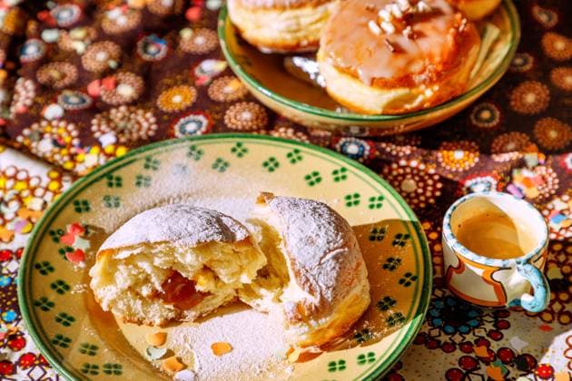 mit Marillenmarmelade gefüllter Faschingskrapfen mit Puderzucker, in der Mitte zerteilt, Keramikteller weiß-grün melliert, Espressotasse, Keramikschale mit verschiedenen Krapfen, gemusterte Tischdecke, Konfetti *** Carnival doughnut filled with apricot jam and powdered sugar, cut in the middle, ceramic plate white green mottled, espresso cup, ceramic bowl with various doughnuts, patterned tablecloth, confetti