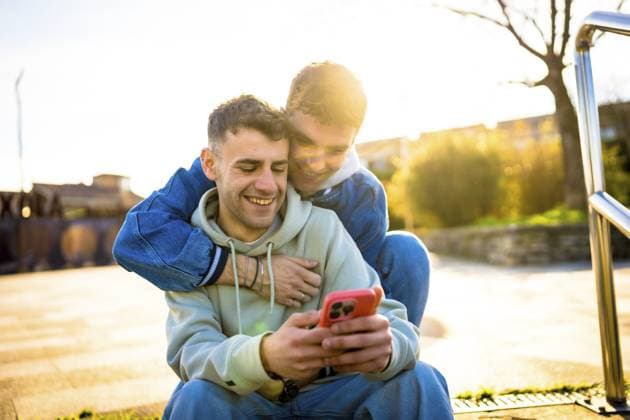 Zwei Männer, die sich umarmen und zusammen lachen, während sie an einem sonnigen Tag im Freien ein Smartphone benutzen, das Freude und Zuneigung ausstrahlt Two men embracing and laughing together while enjoying a smartphone outdoors on a sunny day, radiating joy and affection Copyright: imageBROKER UnaixHuizi ibxuhu12770830.jpg ,model released, Symbolfoto ,property released