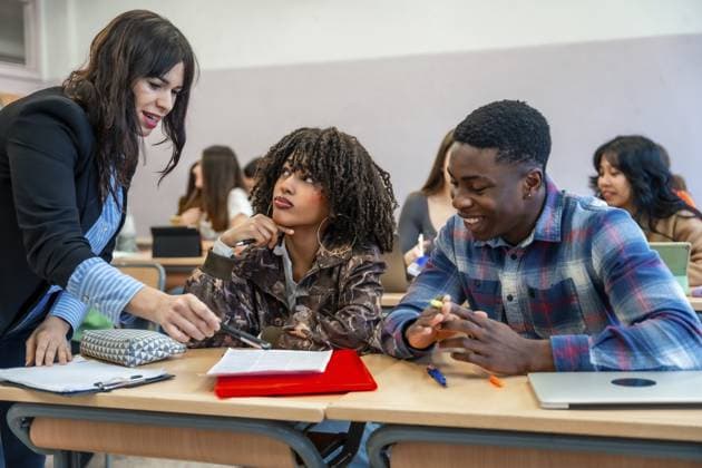 Professor erklärt verschiedenen Universitätsstudenten am Schreibtisch sitzend den Unterricht und hilft ihnen bei ihren Studien Professor explaining lesson to diverse university students sitting at desk, helping them with their studies Copyright: imageBROKER UnaixHuizi ibxuhu12778390.jpg ,model released, Symbolfoto ,property released