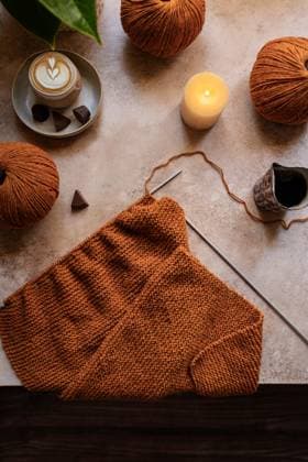 A cozy knitting setup featuring warm brown yarn balls, a knitting project, and a steaming cup of coffee with chocolates, illuminated by a soft candlelight glow. Model Released CintiaViudez_ID26646_768866_002 Copyright: xCintiaxViudezx RECORD DATE NOT STATED