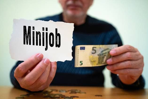 Munich, Bavaria, Germany - February 27, 2025: PHOTOMONTAGE. A man holds a sign saying Minijob and a 5-euro bill in his hand. Mini-jobs play an important role for low-income earners and pensioners who need to supplement their income. *** FOTOMONTAGE. Ein Mann hält ein Schild mit der Aufschrift Minijob sowie einen 5-Euro-Schein in der Hand. Minijobs spielen eine wichtige Rolle für Geringverdiener und Rentner, die ihr Einkommen aufbessern müssen.