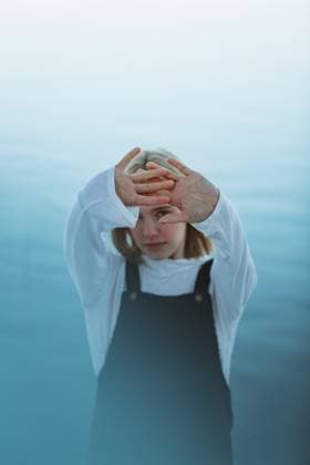 Creative Highlights Symbolfotos A young person in a white shirt and dark overalls stands with arms raised against a serene blue backdrop, expressing a blend of emotion and introspection. Model Released CamilleGirard_ID26913_776697_028 Copyright: xCamillexGirardx