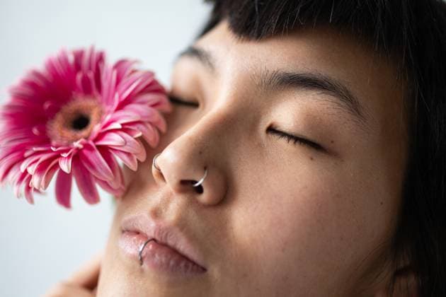 Creative Highlights Symbolfotos Close-up portrait of an Asian woman with closed eyes. She holds a pink flower near her face, showcasing piercings on her nose and lip, conveying a serene expression. Model Released Gaorstudio_ID26044_751628_009 Copyright: xGaorstudiox RECORD DATE NOT STATED