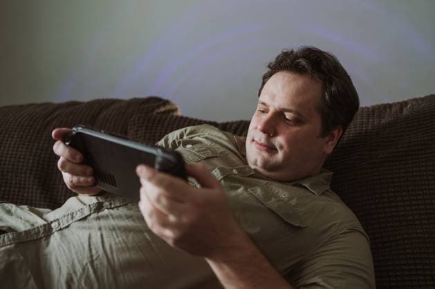 IMAGO Creative, Lifestyle, Gaming A man relaxes on a comfortable couch, focused on playing a game on a hand held device. Casual attire and soft lighting create a cozy home atmosphere. Model Released JenyKulikova_ID26998_779230_022 Copyright: xJenyxKulikovax