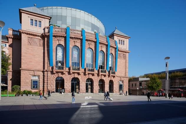 Menschen gehen vor dem Mainzer Staatstheater spazieren und genießen einen klaren und hellen Tag.