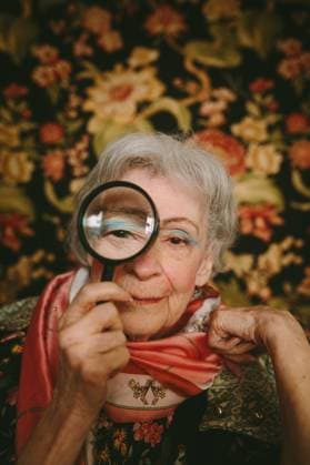 Creative Highlights Symbolfotos An elderly woman holds a magnifying glass to her eye, set against a vibrant floral backdrop. Her vintage clothing and friendly expression create an inviting, warm ambiance. Model Released CamilleGirard_ID26984_778888_003 Copyright: xCamillexGirardx RECORD DATE NOT STATED