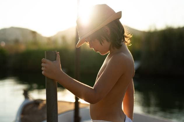 Creative Highlights Symbolfotos Side view of a young boy wearing a straw hat is captured leaning on a boat railing, looking down at the waters of Andratx, Mallorca. The warm sunset creates a serene backdrop emphasizing a peaceful maritime scene. Model Released MagdalenaCarli_ID27192_782031_001 Copyright: xMagdalenaxCarlix