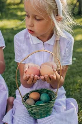 IMAGO Creative, Ostern, Bräuche, Eiersuche #2 A young girl in a purple dress holds Easter eggs with a basket of more colorful eggs on her lap, sitting on grassy lawn. Sunlight enhances the playful scene. Model Released Nastyab_ID27533_790592_035 Copyright: xNastyabx RECORD DATE NOT STATED