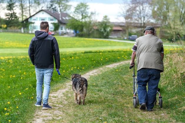 IMAGO Creative, Ostern, Bräuche, Osterspaziergang #2 Bavaria, Germany - April 20, 2025: Two generations out for a walk. A young man leads the dog, an older man walks with a rollator along a meadow path on a spring day *** Zwei Generationen beim Spaziergang. Ein junger Mann führt den Hund, ein älterer Mann läuft mit Rollator über einen Wiesenweg an einem Frühlingstag