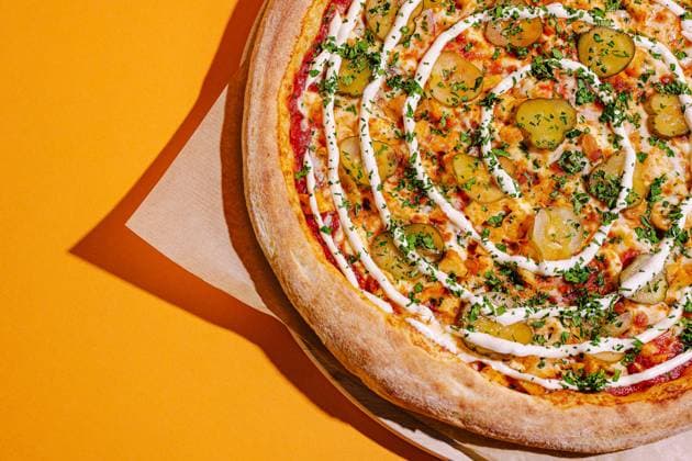 Einzigartige Pizza, belegt mit Essiggurken und beträufelt mit Sauce auf einem leuchtend orangefarbenen Hintergrund A large pizza features a spiral pattern of creamy sauce and vibrant green herbs, with slices of pickles scattered across the cheesy surface, all against a bright orange backdrop. (License=RF) Copyright: xZoonar.com AntonxDobreax 23366683