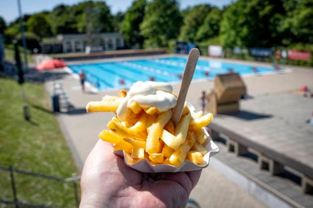 Freibadpommes Frische Pommes mit Mayonnaise in einer typischen weißen Pappschale werden von einer Hand in den Vordergrund gehalten, während im Hintergrund ein sonniges Freibad mit blauem Schwimmbecken und Badegästen zu sehen ist. Die Szene vermittelt Sommergefühle, Freizeitspaß und Genuss im Freien. Die Pommes sind goldgelb frittiert, mit einem Klecks Mayonnaise garniert und einem kleinen Holzpiekser serviert ein klassischer Snack in deutschen Freibädern. Die Atmosphäre ist entspannt, das Wetter sonnig, und das Freibad von grünen Bäumen umgeben. Veröffentlichungen nur für redaktionelle Zwecke. Foto: FotoPrensa Essen *** Outdoor pool fries Fresh fries with mayonnaise in a typical white cardboard bowl are held by a hand in t