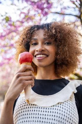 Lächelnde junge Frau, die eine Eiswaffel genießt, während sie unter einem blühenden Baum steht, umgeben von leuchtenden Blumen an einem sonnigen Frühlingstag Smiling young woman enjoying an ice cream cone while standing under a blooming tree, surrounded by vibrant flowers on a sunny spring day Copyright: imageBROKER UnaixHuizi ibxuhu13695103.jpg ,model released, Symbolfoto ,property released