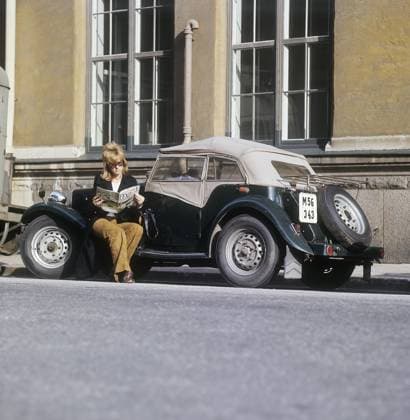 Eine Frau neben einem Sportwagen. Das Auto ist ein englisches MG TD Modell, das zwischen 1950 und 1953 hergestellt wurde. Dieses Modell war besonders gut für schwedische Kunden geeignet, da es mit dem Lenkrad auf der linken Seite gebaut werden konnte, was den damaligen schwedischen Fahrgewohnheiten entsprach. Insgesamt wurden 29.664 Einheiten des MG TD hergestellt. Sie sitzt auf dem Wagen und liest die Frauenzeitschrift Femina. Sie ist mit einem Blazer und einer braunen Hose bekleidet. 1960er Jahre Schweden. Weitere Fakten zum MG TD Der MG TD, Teil der MG T Type Serie, war ein zweisitziger Roadster, der für sein klassisches britisches Design bekannt war, mit einem markanten aufrechten Kühlergrill, freiliegenden Rädern und einer kompakten, Copyright: imageBROKER Sjoberg ibljsj13777496.jpg