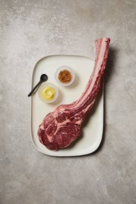 Top-down view of a raw tomahawk steak presented on a ceramic tray, accompanied by a small bowl of mustard, spices, and a black spoon. A premium cut ready for gourmet cooking Model Released GeorgiaGold_ID28577_843647_038 Copyright: xGeorgiaxGoldx